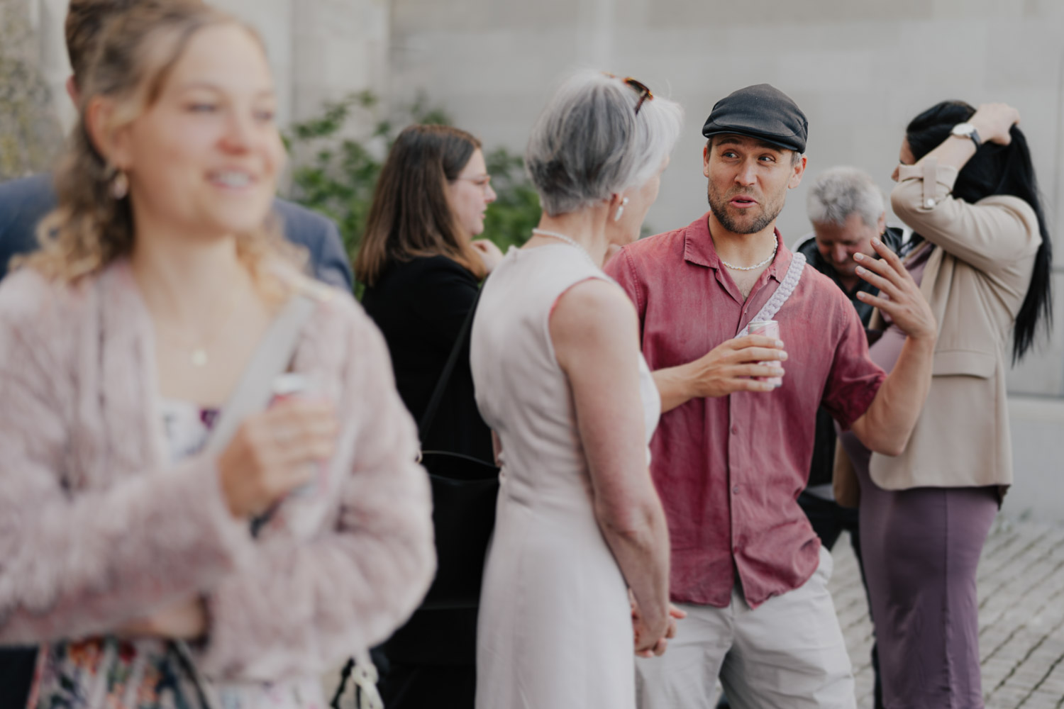A group of people socializing outdoors. A man in a pink shirt and cap gestures while speaking to a woman with gray hair. Captured by a Hochzeitsfotograf Zürich, others chat in the background, with one woman smiling in the foreground.