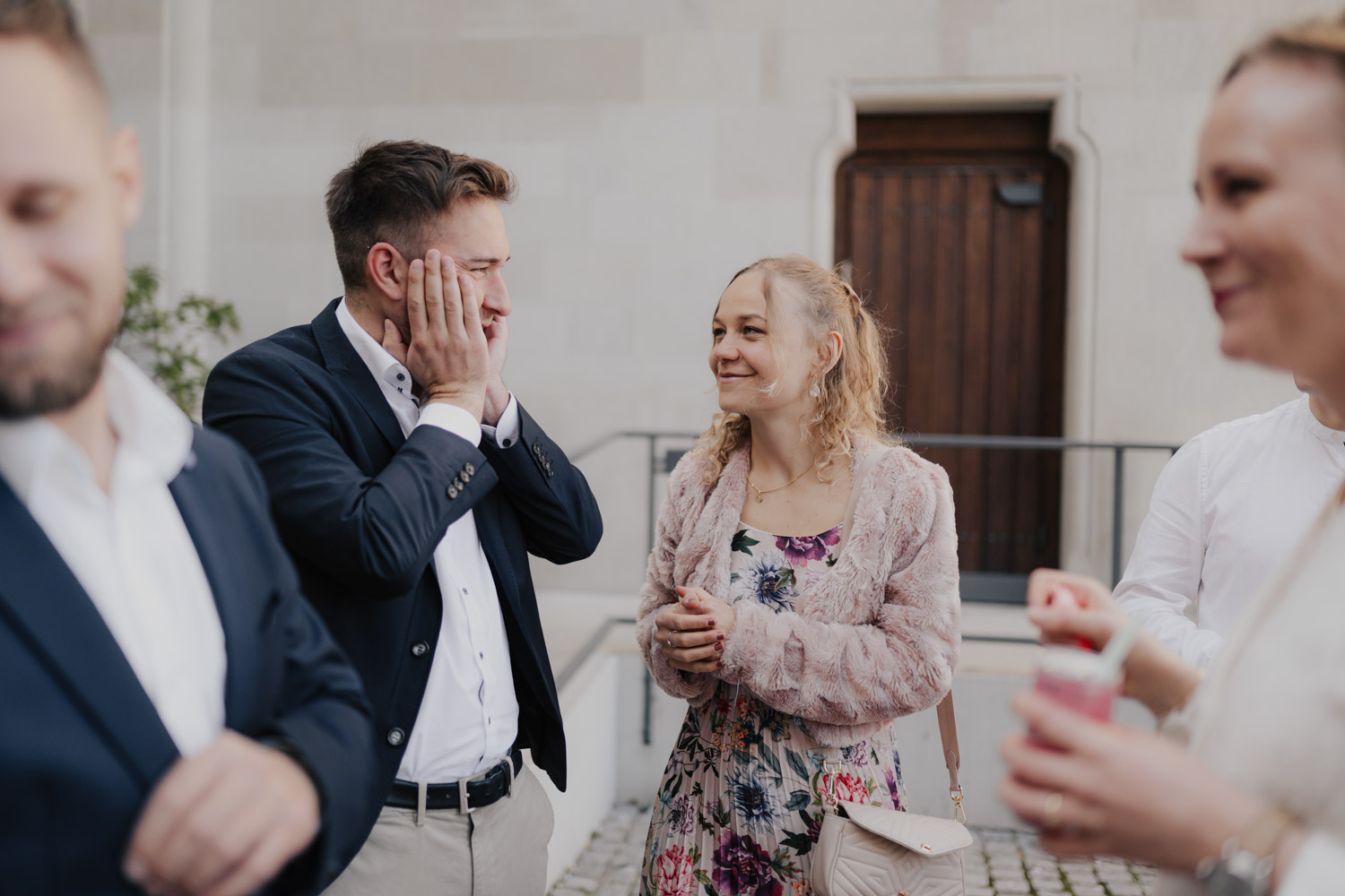 A man in a navy blazer holds his face in surprise while a smiling woman in a floral dress and pink jacket looks on. Two others stand nearby, all outdoors before a stone building—captured by Hochzeitsfotograf Zürich.