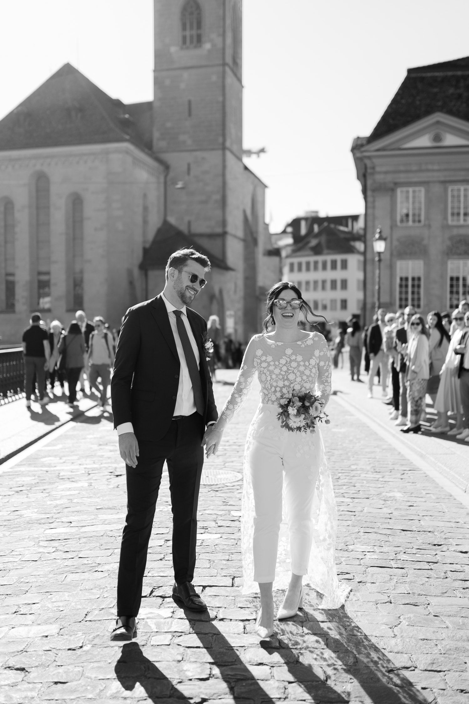 A newlywed couple, smiling and holding hands, walks on a sunlit cobblestone street. Captured by a Hochzeitsfotograf Zürich, the bride in glasses and the groom in a suit delight as people watch near historic buildings.