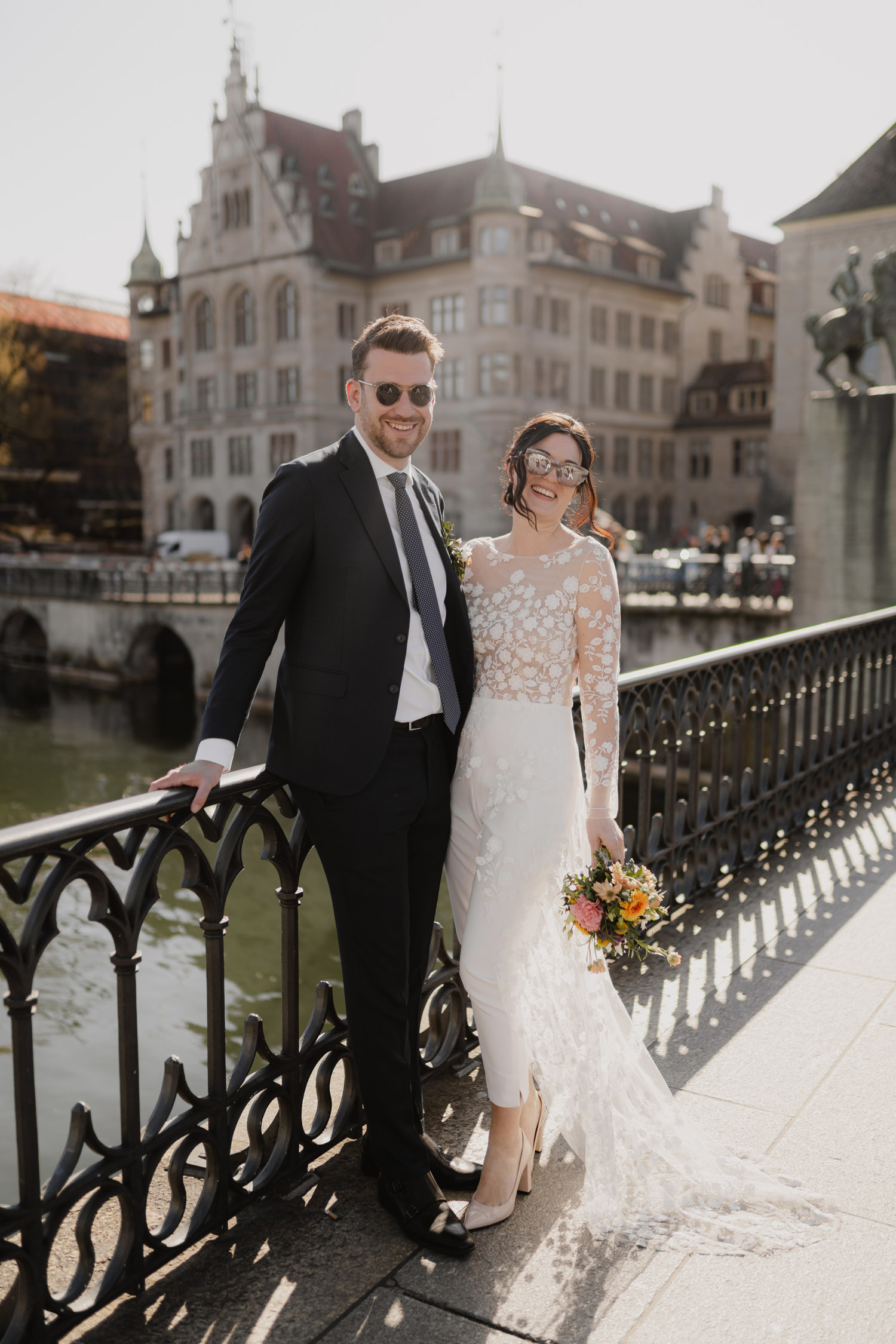 A couple dressed in wedding attire, both wearing sunglasses, stand smiling on a bridge with historic buildings and water in the background. Captured by a Hochzeitsfotograf Zürich, the bride holds a bouquet and wears a lace dress with sheer sleeves.