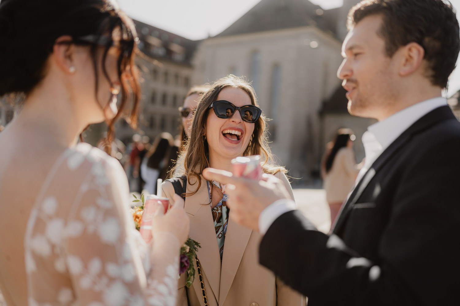 Three people stand outdoors, laughing and talking. A woman in sunglasses and a beige coat joins a formally dressed couple with drinks. Sunlight brightens the scene, captured by Hochzeitsfotograf Zürich, with a building in the background.