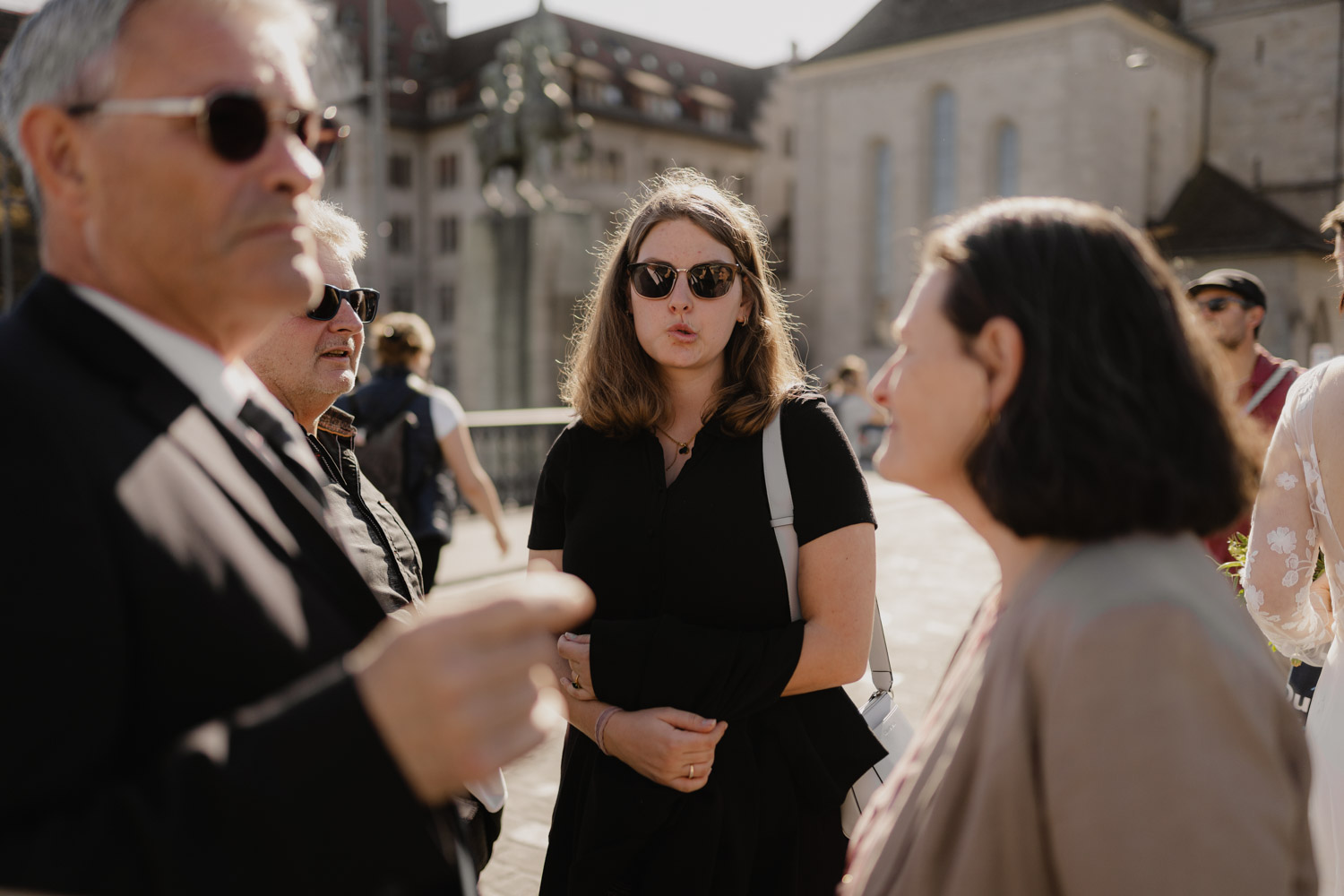 Three people wearing sunglasses stand outdoors in sunlight, engaged in conversation. A woman in black stands at the center with her arms crossed, while two others face her—perfect inspiration for any Hochzeitsfotograf Zürich capturing candid moments.