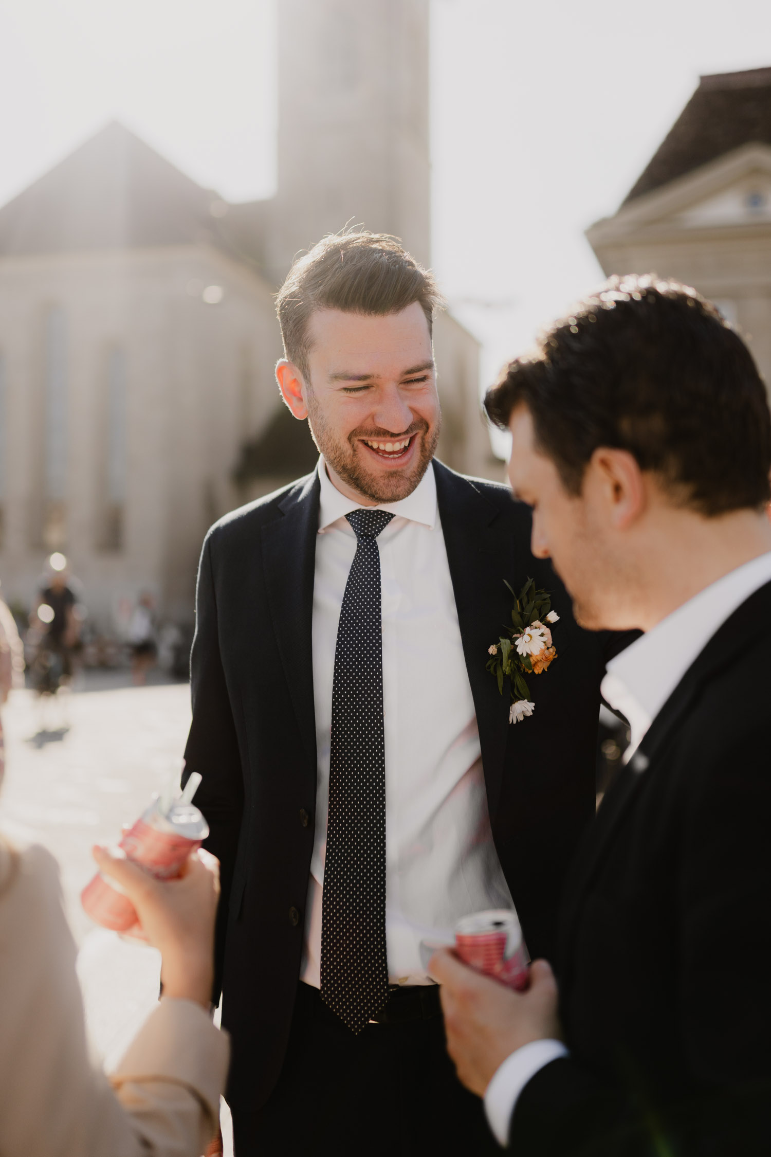 A man in a suit with a boutonnière smiles and chats with two people outdoors in sunlight. They’re dressed formally, holding drinks, with buildings blurred behind—captured by a Hochzeitsfotograf Zürich for a stylish Swiss wedding moment.