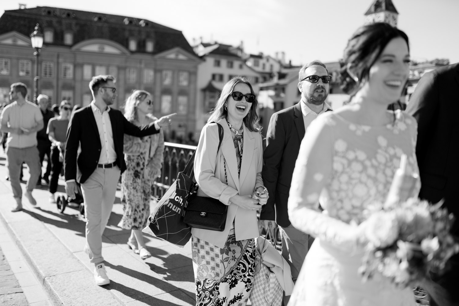 A group of well-dressed people walk outdoors in sunlight, smiling and laughing. Captured by a Hochzeitsfotograf Zürich, the focus is on a woman in sunglasses and patterned skirt; a bride in white holding flowers is blurred in the foreground.