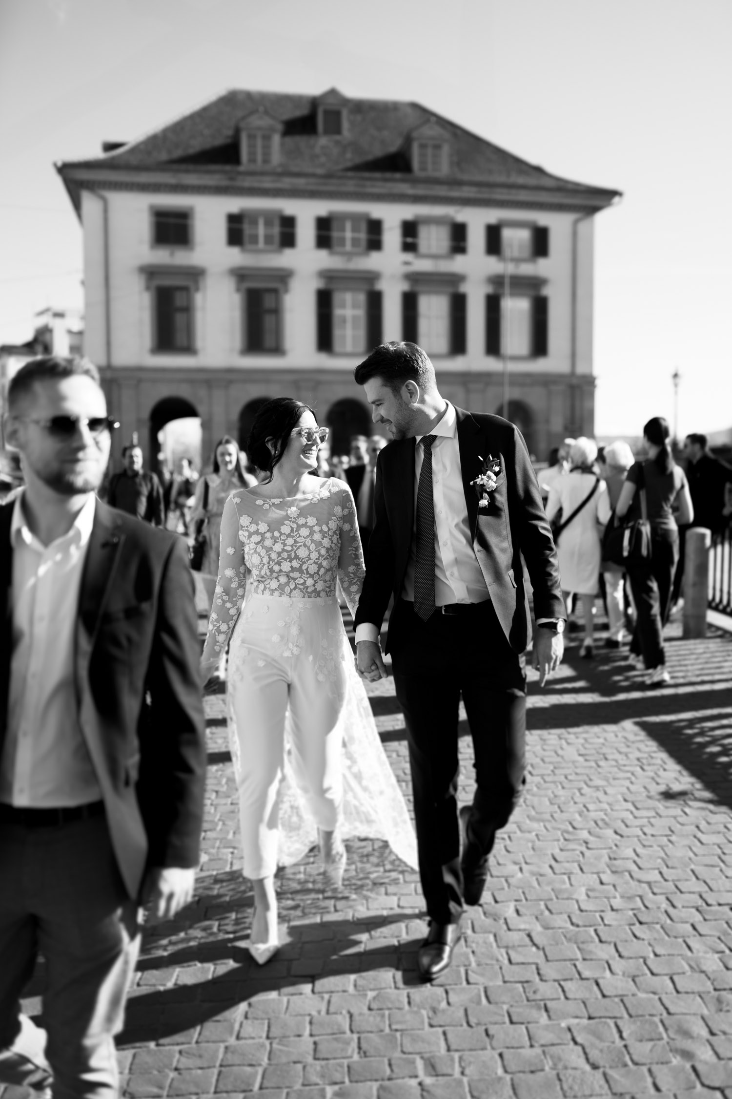 A bride and groom walk hand in hand, smiling at each other, surrounded by people on a cobblestone street in front of a large building. This timeless black and white moment was beautifully captured by a Hochzeitsfotograf Zürich.