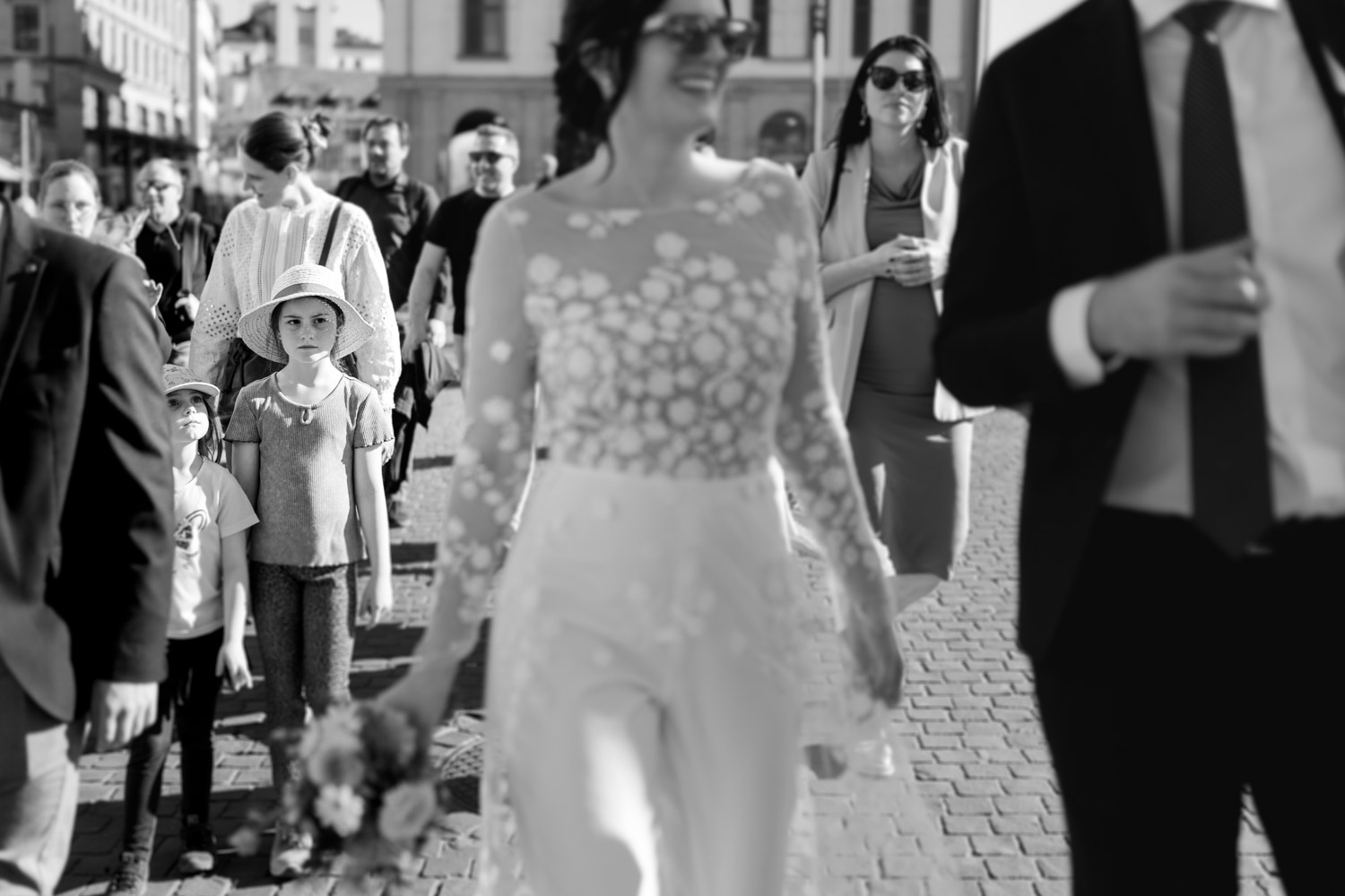 A group walks outdoors on a sunny day. In focus, a serious young girl in a hat stands among smiling adults, including a bride in a floral dress and bouquet, captured by a Wedding Photographer Zurich, with adults out of focus in the foreground.